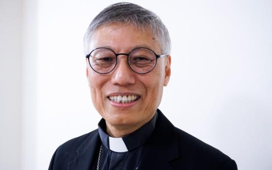 An East Asian man wearing round glasses and a clerical collar smiles at the camera