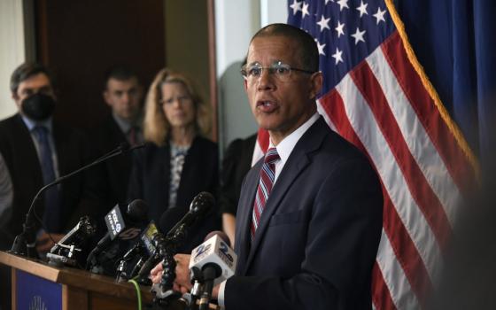 A light-skinned Black man with glasses in a suit speaks from behind a podium with many news microphones