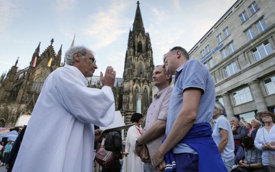An older white man in white vestments holds his hand as though making the sign of the cross in front of two white men