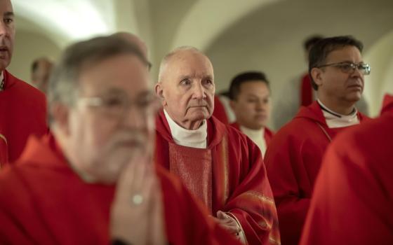 An older white man wearing red vestments stands among other men wearing red vestments