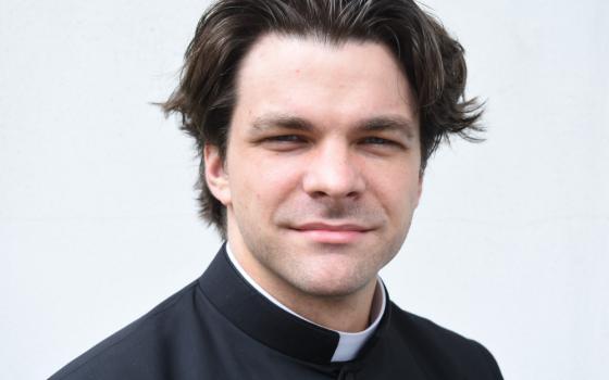 A white man wearing a clerical collar with styled, swoopy hair looks into the camera