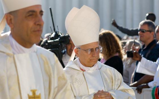 Two light-skinned men wear white mitres and vestments as they stand side-by-side with people and cameras in the background