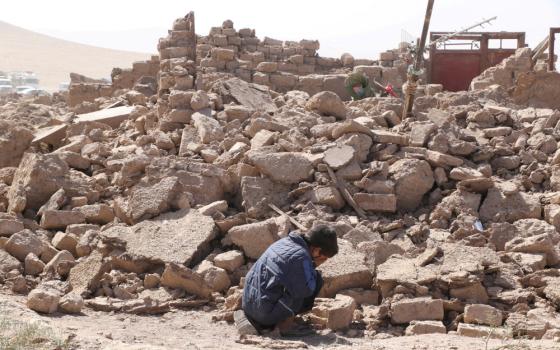 A boy wearing a blue jacket crouches with his face in his hands beside a large pile of rubble