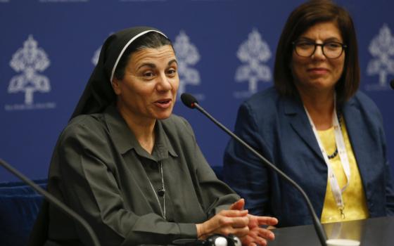 A brown nun speaks into a microphone while a brown woman in Western business clothes sits beside her