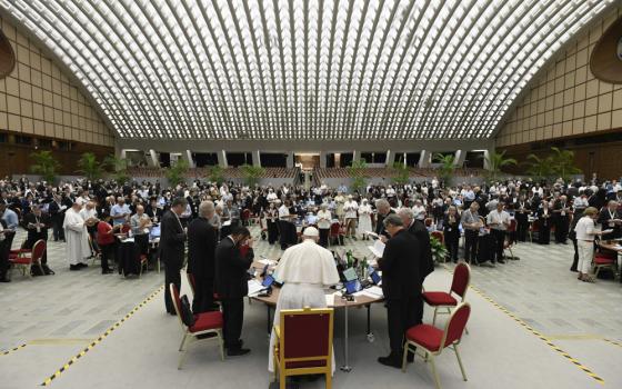 Many people stand around many round tables in a domed hall. Pope Francis is visible at a table in the front.