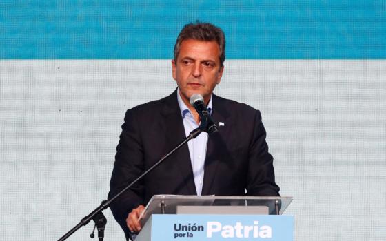 A light-skinned man with black and gray hair who is wearing a suit jacket speaks into a microphone at a podium that says "Union por la patria"