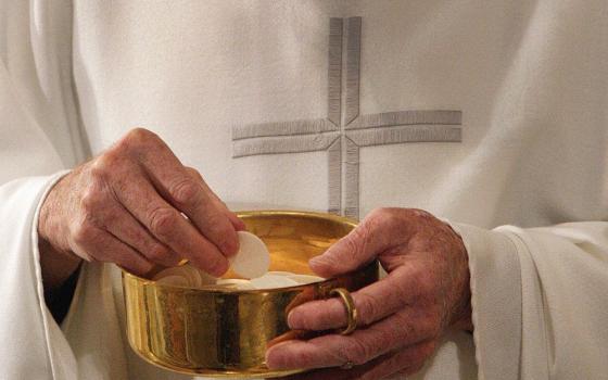 A white man's hands hold a pyx with Communion wafers. The man wears a white vestment with a cross on it.