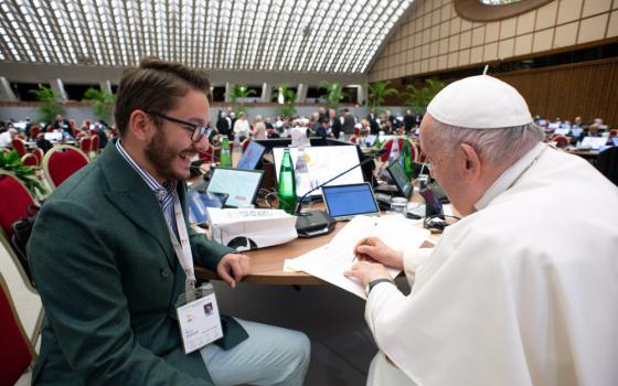 A young man with brown hair wearing a suit smiles at Pope Francis as he signs a document