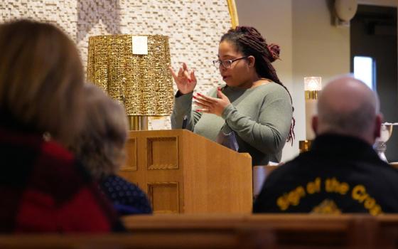 A Black woman wearing glasses signs from behind a wooden lectern. The backs of parishioners in pews watching her are visible.