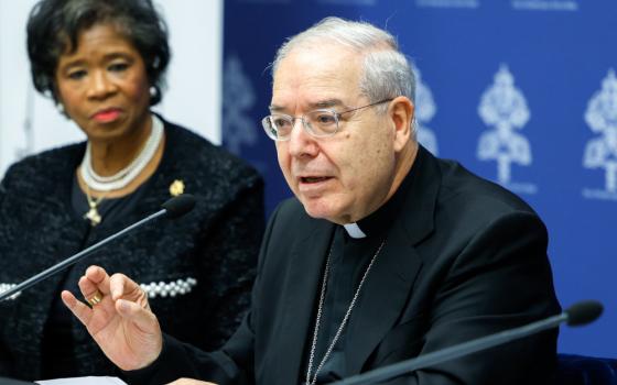 An older white man with glasses wearing a clerical collar speaks into a microphone while a professionally dressed older Black woman looks on