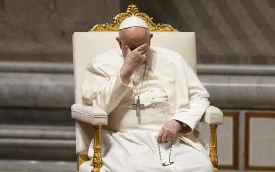 Pope Francis places one hand over his eyes and holds his rosary in the other while seated in a white and gold chair.