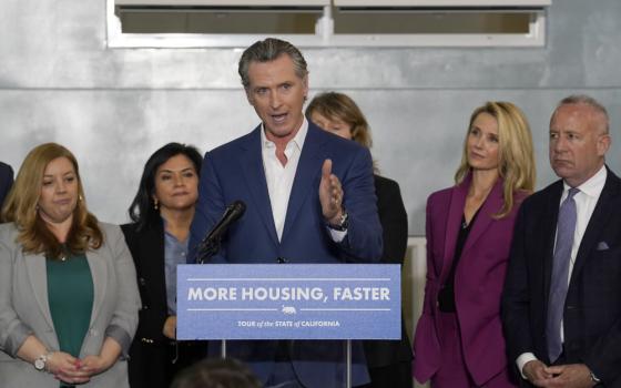 A white man with graying hair wearing suit jacket with no tie speaks from behind a podium that reads "more housing, faster" as others in business professional wear stand behind him