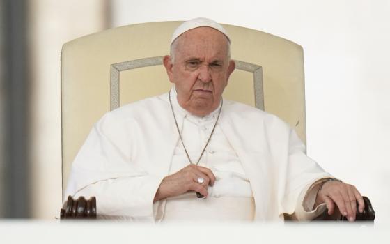 Pope Francis sits in his white chair with a very serious face as his hands close around his pectoral cross