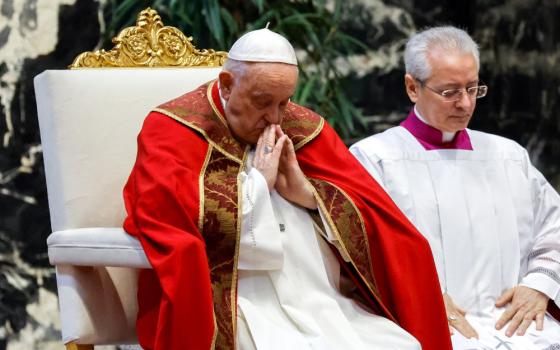 Pope Francis wearing a red cape over his ususal white cassock bows his head and holds his palms together at his chest while sitting in a white and gold chair.