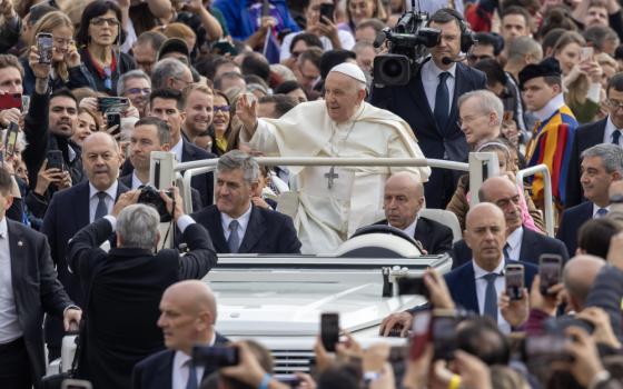 Pope Francis waves at the crowd surrounding his popemobile. A TV camera films him in the popemobile.