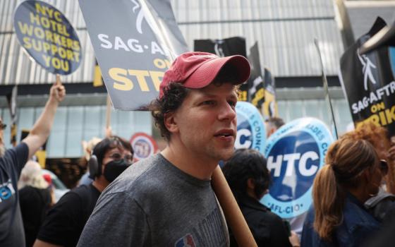A white man wearing a ball cap has a SAG-AFTRA on strike sign over his shoulder while surrounded by other people with signs outside an office building