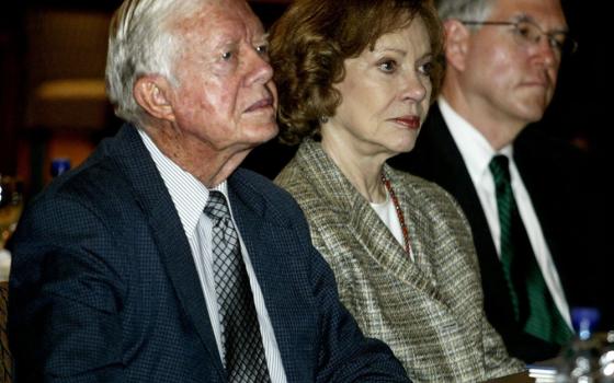 A brown-haired older white woman sits between two older white men. All wear formal clothes.
