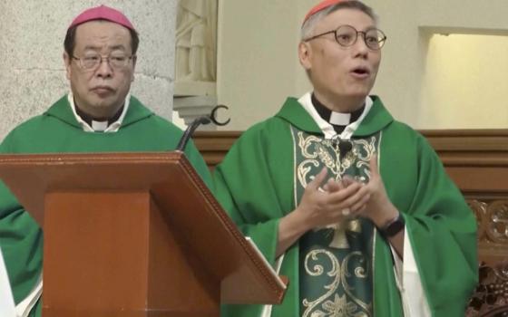 An East Asian man wearing green vestments and a purple zucchetto stands next to another east Asian man wearing green vestments and a red zucchetto. They both stand behind a wooden podium.