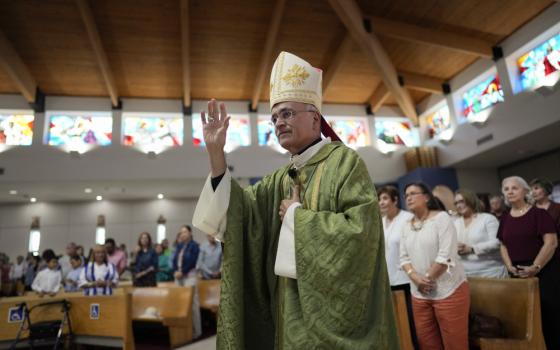 A man wearing green vestments and a white mitre raises his right hand while standing before a crowded church