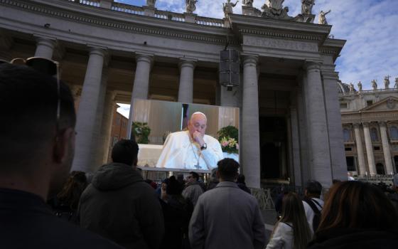 Pope Francis, sitting and holding a fist towards his chin, is projected on a large screen in front of a crowd in St. Peter's Square