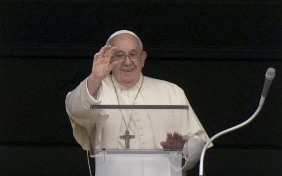 Pope Francis raises his right hand and smiles as he stands behind a clear lectern