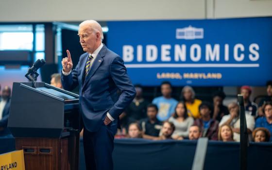 President Biden stands at lectern with people watching him and sign behind him reading "Bidenomics."