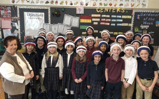 Woman stands with a group of children in a classroom. The children are wearing crocheted headbands,