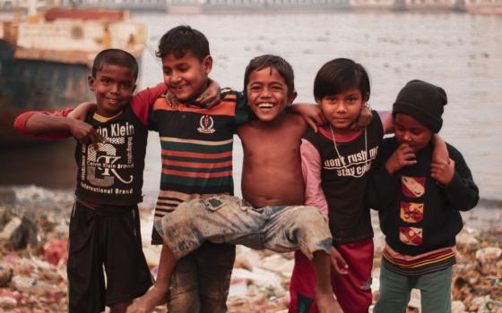 A group of children stands in a line, arms across each other's shoulders. A body of water is behind them. 