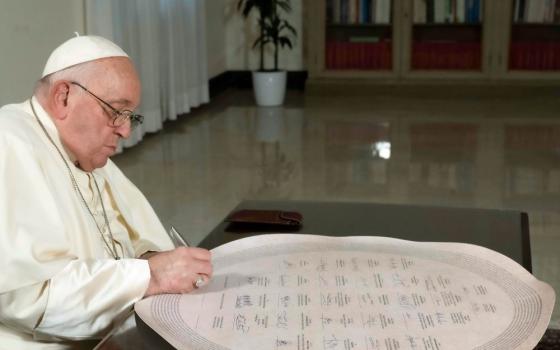 Pope Francis writing on a large, round, white item on a desk.