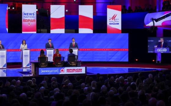 Four presidential candidates on stage for debate, while screen to the right shows former U.S. President Donald Trump