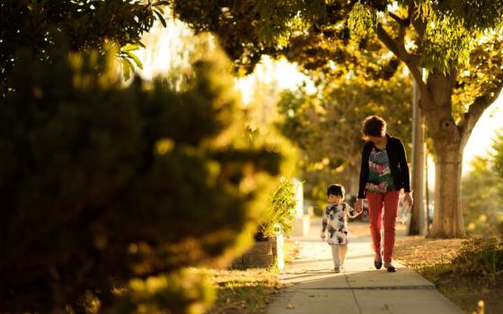 Woman and young boy hold hands and walk along sidewalk surrounded by trees.