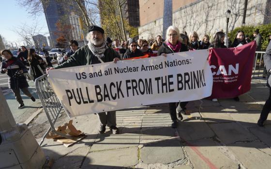 Two people hold a banner that says "U.S. and Nuclear Armed Nations:  Pull Back From The Brink" while walking in front of a crowd