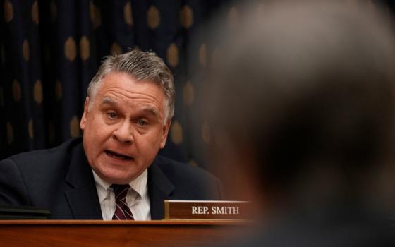 A graying white man wearing a suit and tie speaks next to a Rep. Smith name plate