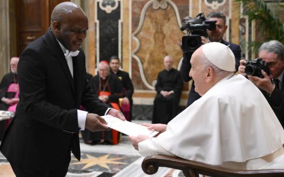A Black man standing and wearing a white-tie tuxedo hands a seated Pope Francis a piece of paper
