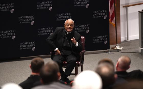 An older Black man wearing a clerical collar and black jacket sits in front of an audience