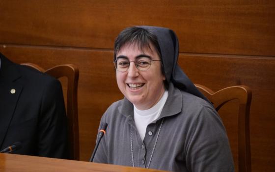 A woman religious wearing glasses and a gray habit sits at a table and speaks into a microphone