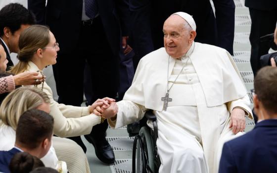 Pope Francis, sitting in a wheelchair, holds the hands of a young woman wearing glasses