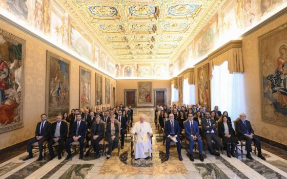 Pope Francis sits in a wheelchair between two sections of seated people in business attire