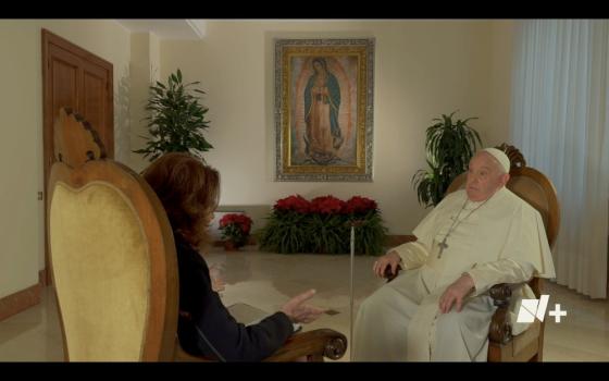 Pope Francis sits in a chair opposite another person in a chair in a room with a framed Virgen de Guadalupe image, poinsettias and plants