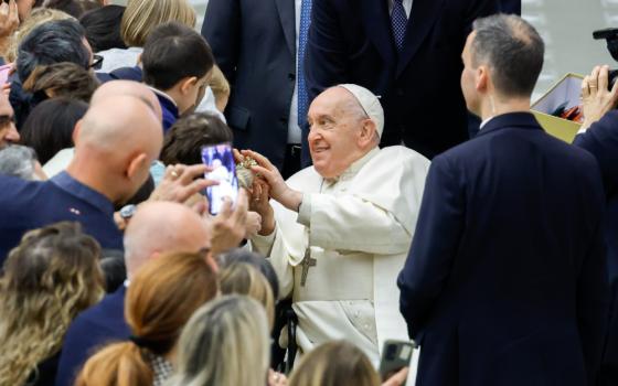 Pope Francis sits in a wheelchair rolling past a crowd of people, turning to the side to rest his hands on an object.