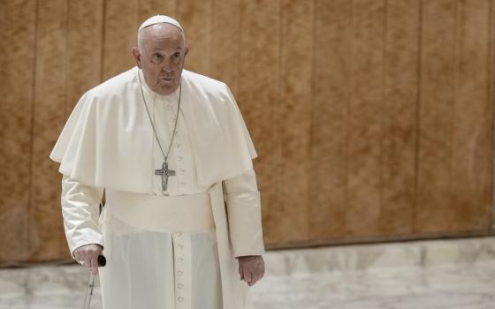 Pope Francis walks with a serious expression with a cane in front of a wooden wall and marble floor