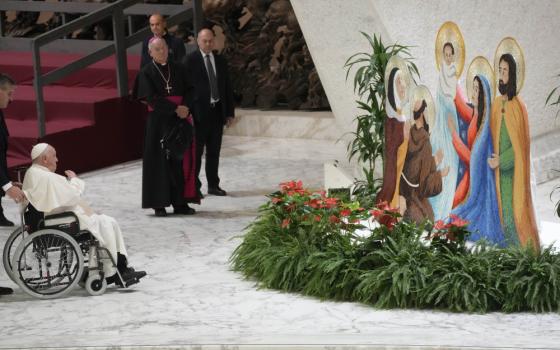 Pope Francis sits in a wheelchair before figures of the Holy Family and greenery