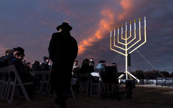 A very large menorah stands in the darkening sky with orange clouds before a crowd