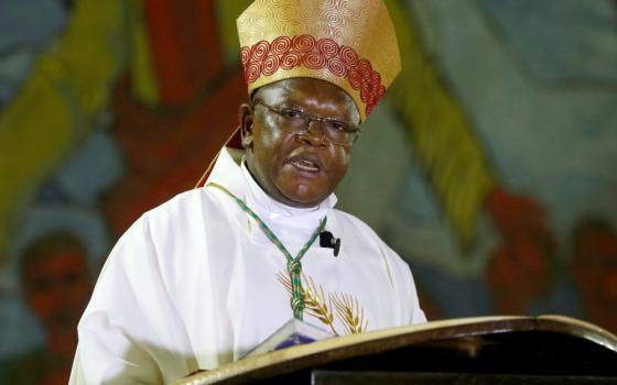 A Black man wearing glasses, a gold and red mitre and white vestments speaks behind a podium