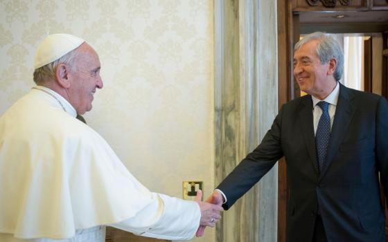 An older white man wearing a suit shakes Pope Francis' hand in a room with white patterned wallpaper