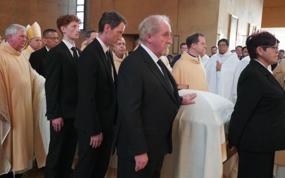 Men in suits and vestments surround a casket draped in a white sheet 