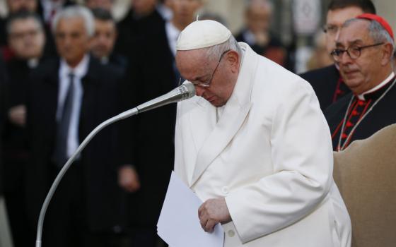 Pope Francis bows his head in front of a microphone. Men in suits and cardinal's cassocks are visible in the background.