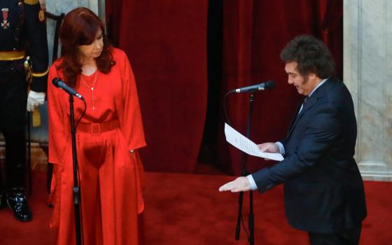 A white man in a suit reads from a paper with his other palm face down while standing next to a woman in a red dress