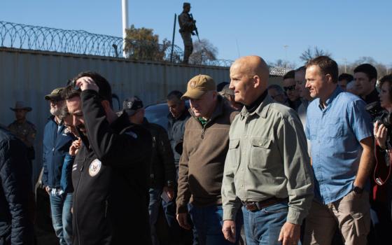 A group of white men in casual clothes walk along a concrete wall topped with barbed wire