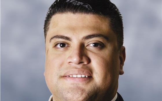 A Latino man with short hair and a clerical collars looks into the camera with a slight smile